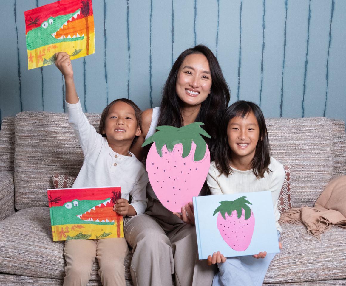 A mom and her two kids on the couch holding up their artwork and the printed Kids Art Book